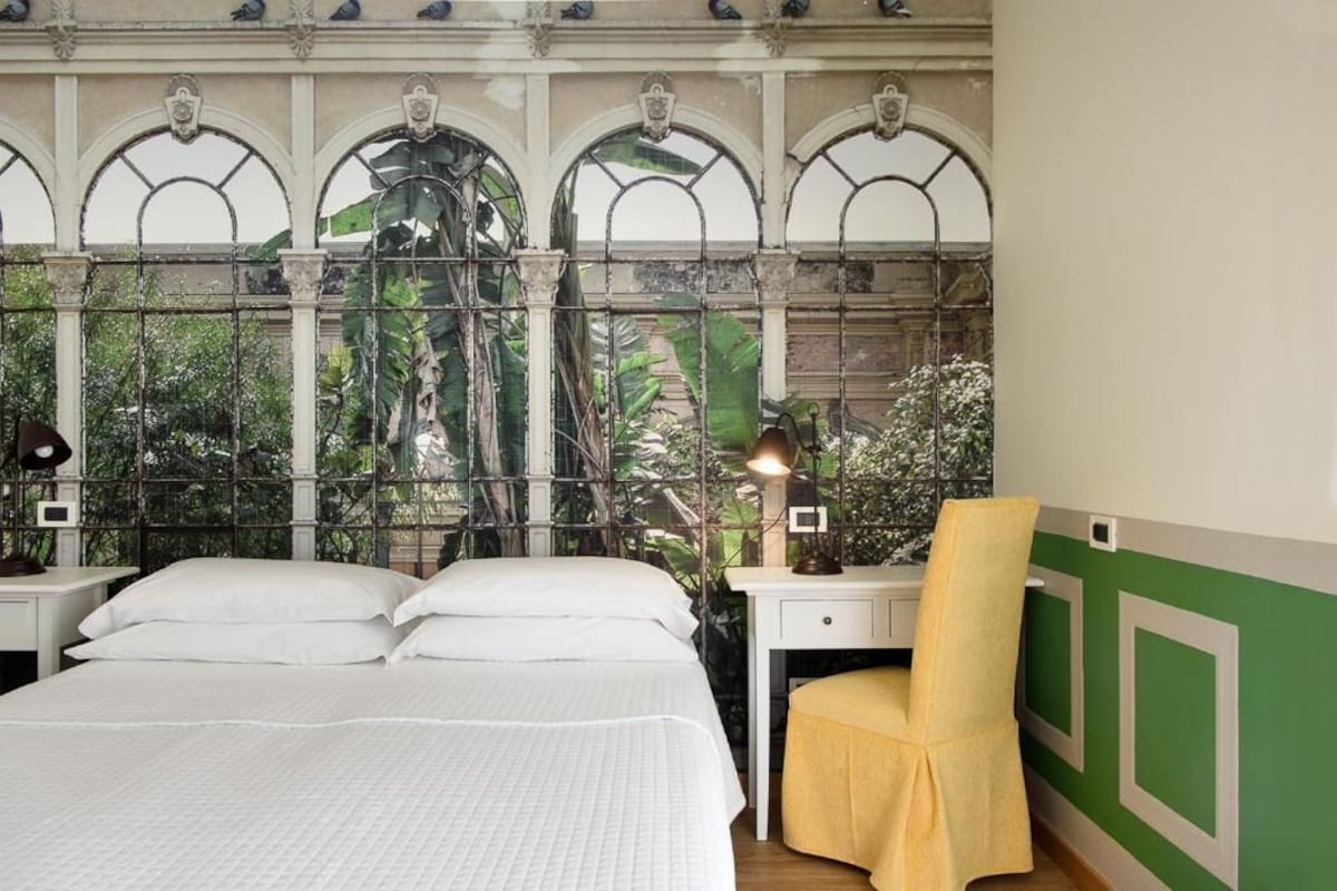 Habitació d'hotel amb un llit, una taula de treball, una cadira i una finestra amb vista a vegetació.