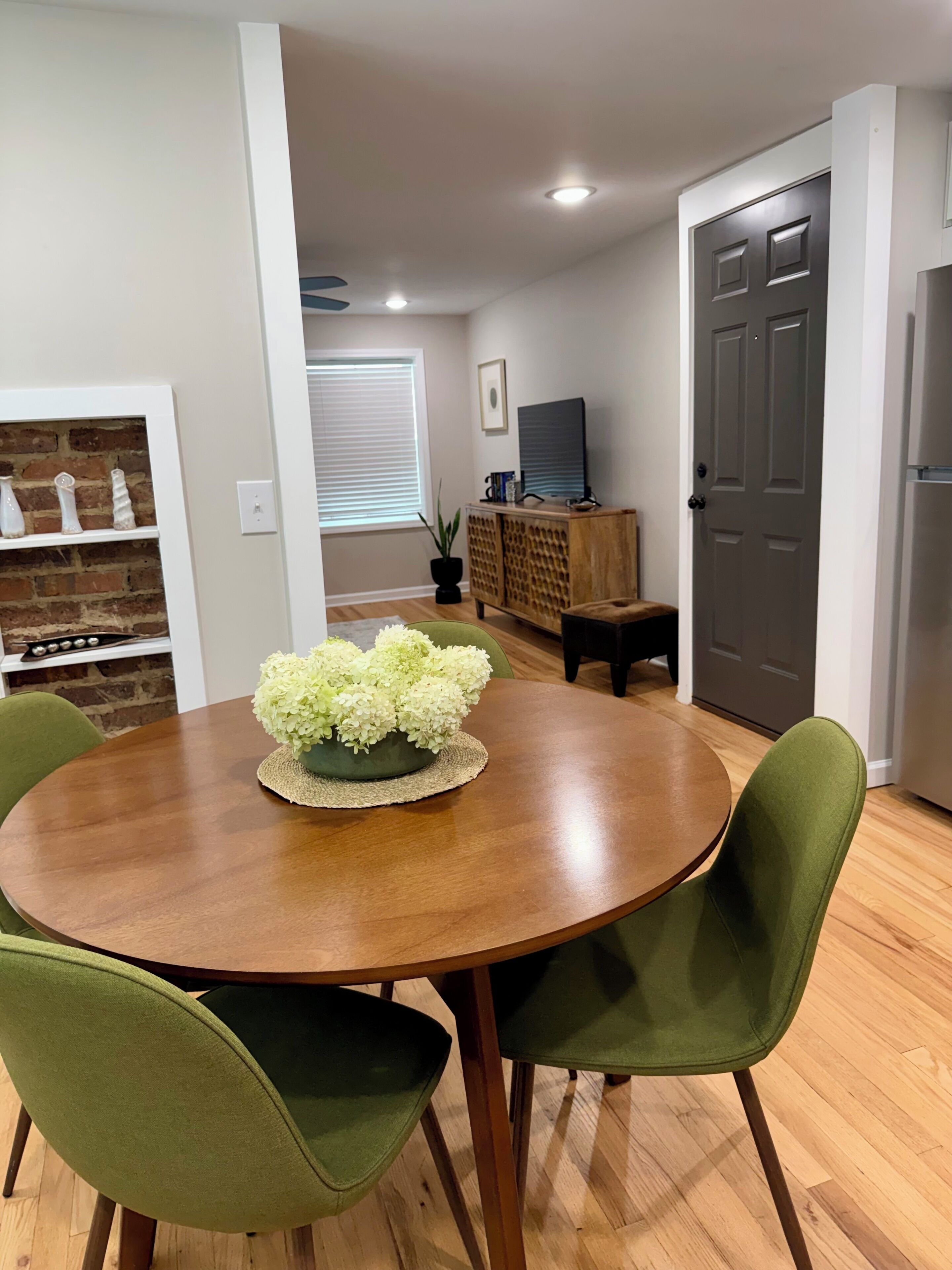 A dining area with a round wooden table, green upholstered chairs, and a centerpiece of white flowers.