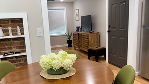 Un área de comedor con una mesa redonda de madera, sillas tapizadas en verde y un centro de mesa con flores blancas.