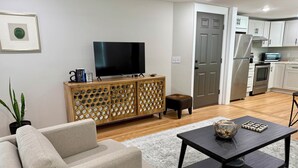 A living room with a sofa, coffee table, TV, wooden cabinet, and a kitchen area with cabinets and appliances.