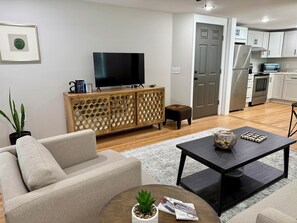 Una sala de estar con sofá, mesa de centro, televisor, mueble de madera y una zona de cocina con armarios y electrodomésticos.