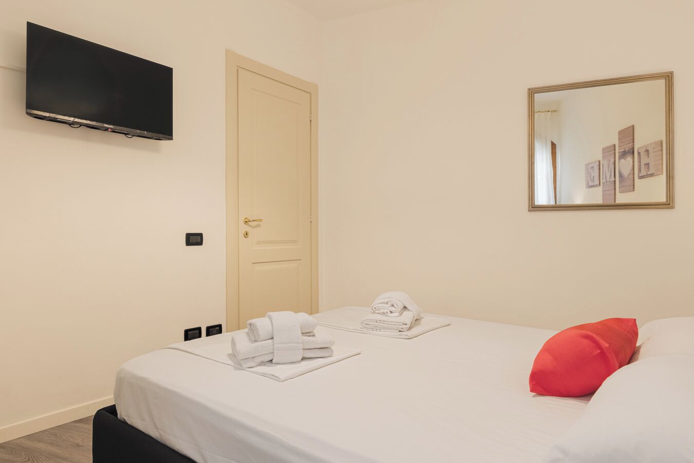 A hotel room with a bed, a television mounted on the wall, a door, and a mirror with a decorative frame.