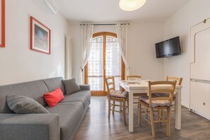 A living room with a grey sofa, a dining table with chairs, a television mounted on the wall, and a window with curtains. - Appartamento Dogana by Wonderful Italy (Peschiera del Garda)