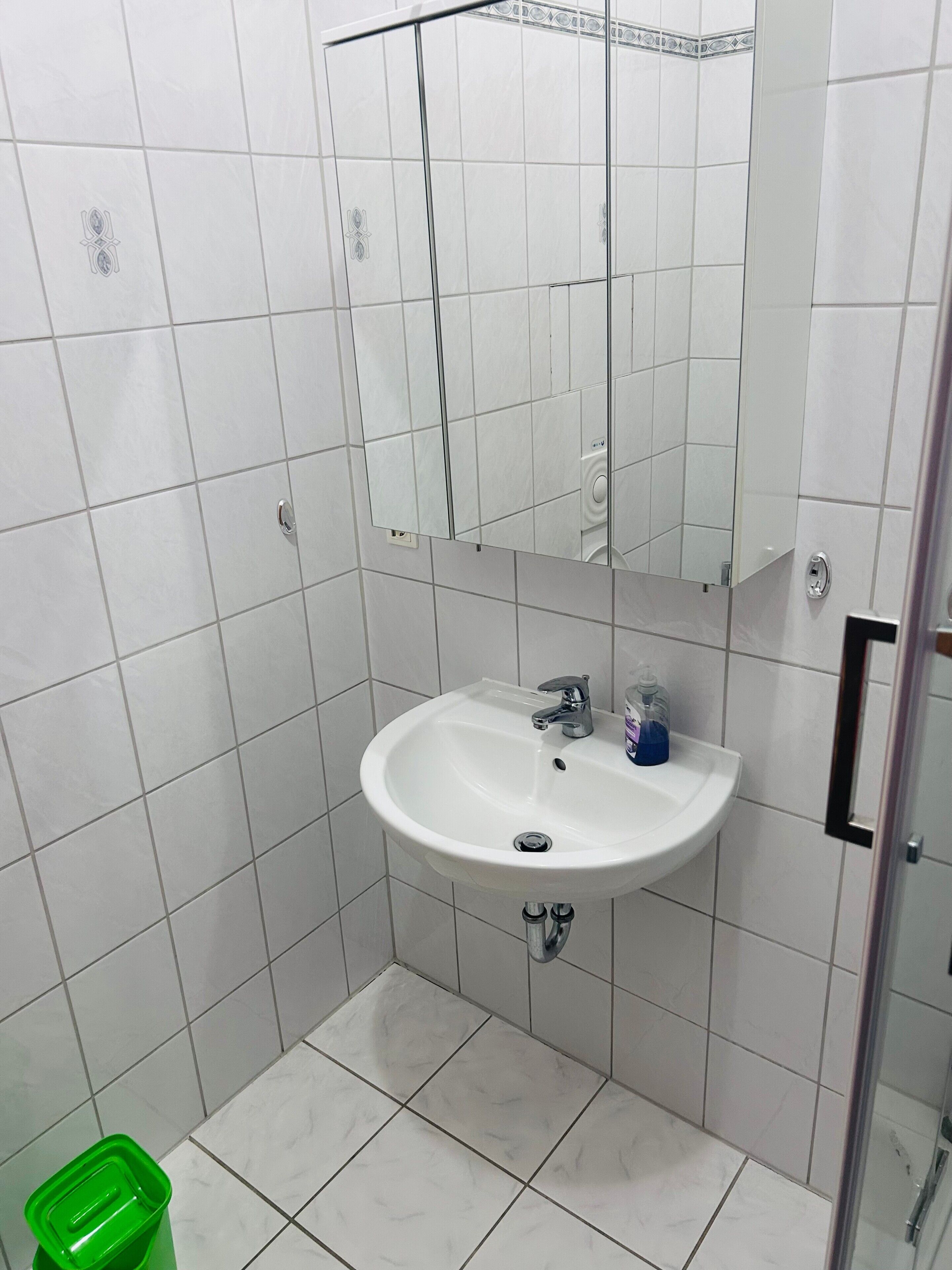 A bathroom with white tiles, a sink, a mirror, and a green trash bin.