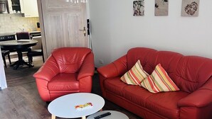 Una sala de estar con un sofá de cuero rojo, dos mesas de centro blancas, una araña y una puerta que da a una cocina.