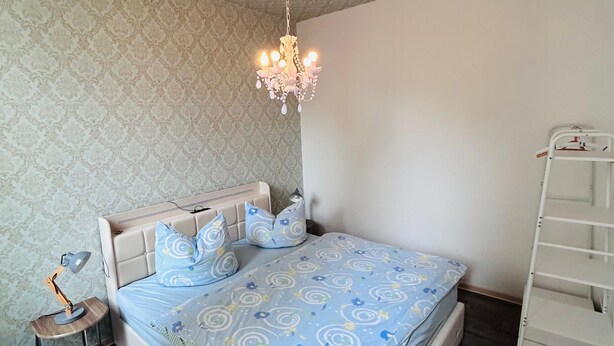 Un dormitorio con cama, lámpara de noche, candelabro y armario blanco.