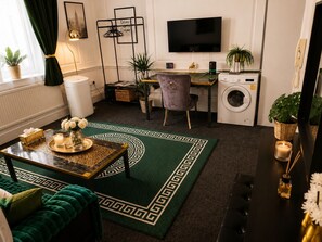 Ein modernes Wohnzimmer mit einem grün gemusterten Teppich, einer Couch, einem Couchtisch mit Dekorationen, einem Fernseher, einer Waschmaschine und einem Schreibtisch mit Stuhl.