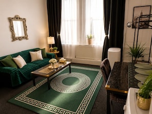 Ein Wohnzimmer mit einem grünen Sofa, einem Couchtisch, einem Esstisch mit Stühlen, einem Spiegel und einem Fenster mit Vorhängen.