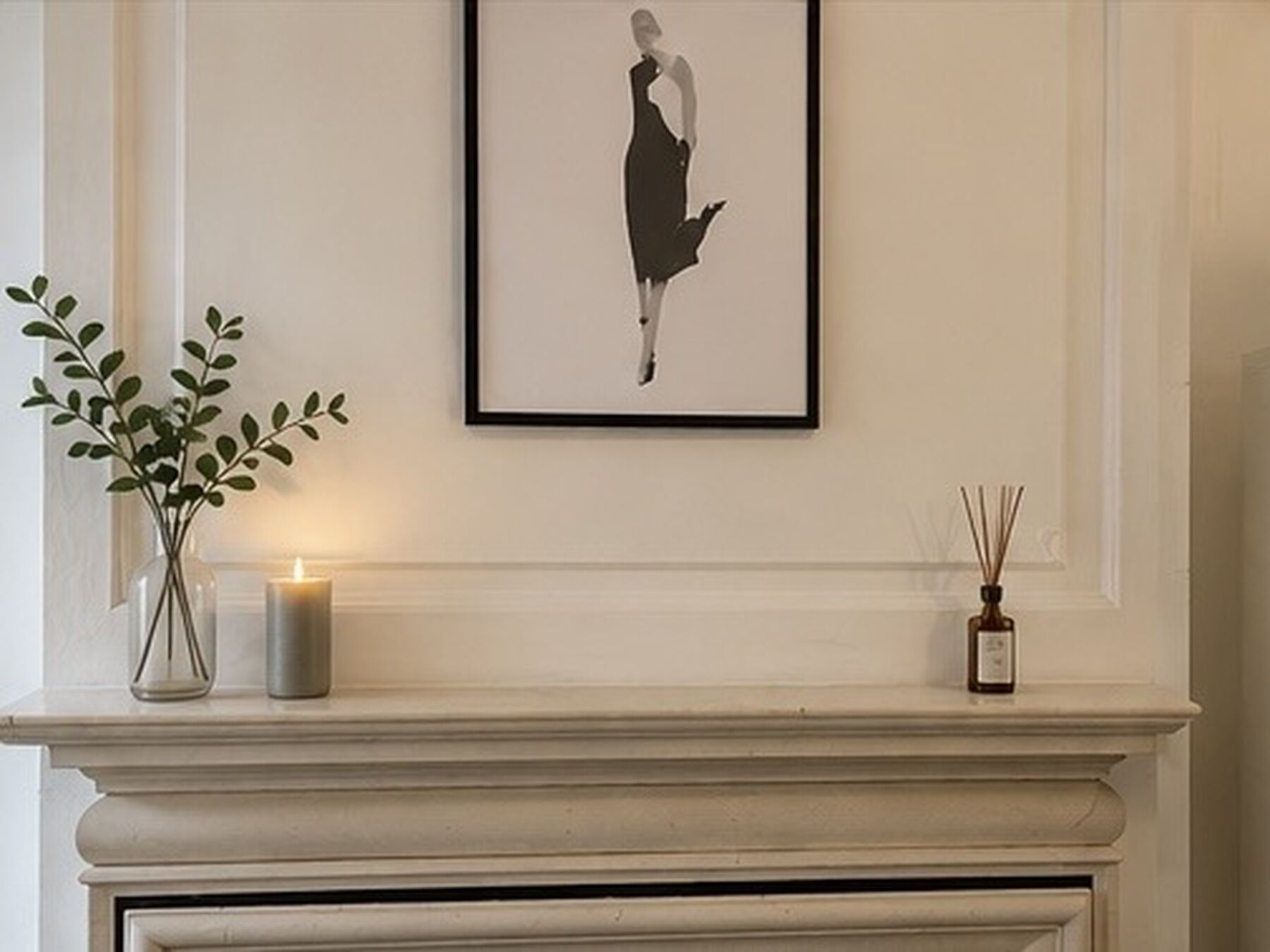 A framed black and white artwork on a wall above a fireplace mantel with a candle and a reed diffuser.