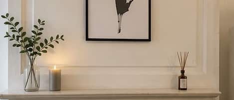 A framed black and white artwork on a wall above a fireplace mantel with a candle and a reed diffuser.