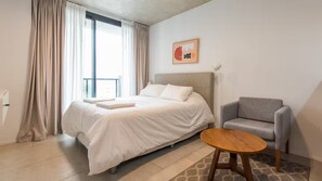 Une chambre d’hôtel avec un lit, un fauteuil gris, une petite table en bois et une fenêtre avec des rideaux.