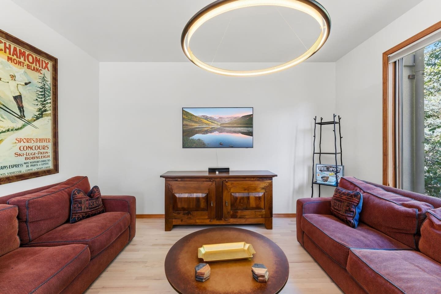 A modern living room with two red sofas, a wooden coffee table, a mounted TV, and a large window.