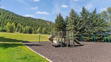 A playground with a slide and climbing structures, surrounded by grass and trees, with a mountain in the background.