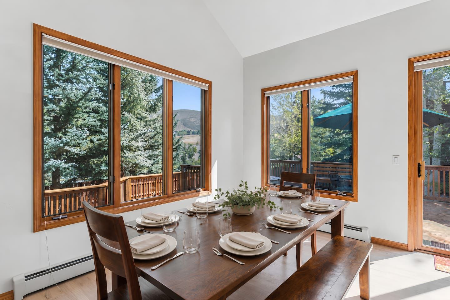 A dining area with a wooden table set for four, large windows with a view of trees, and a balcony with a wooden railing.