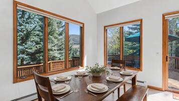 A dining area with a wooden table set for four, large windows with a view of trees, and a balcony with a wooden railing.