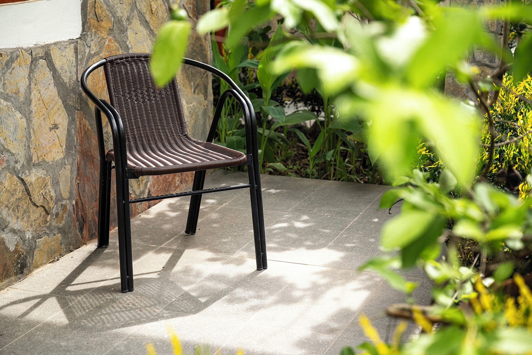 Una silla de mimbre con estructura metálica, ubicada en un patio empedrado con pared de piedra y follaje al fondo.