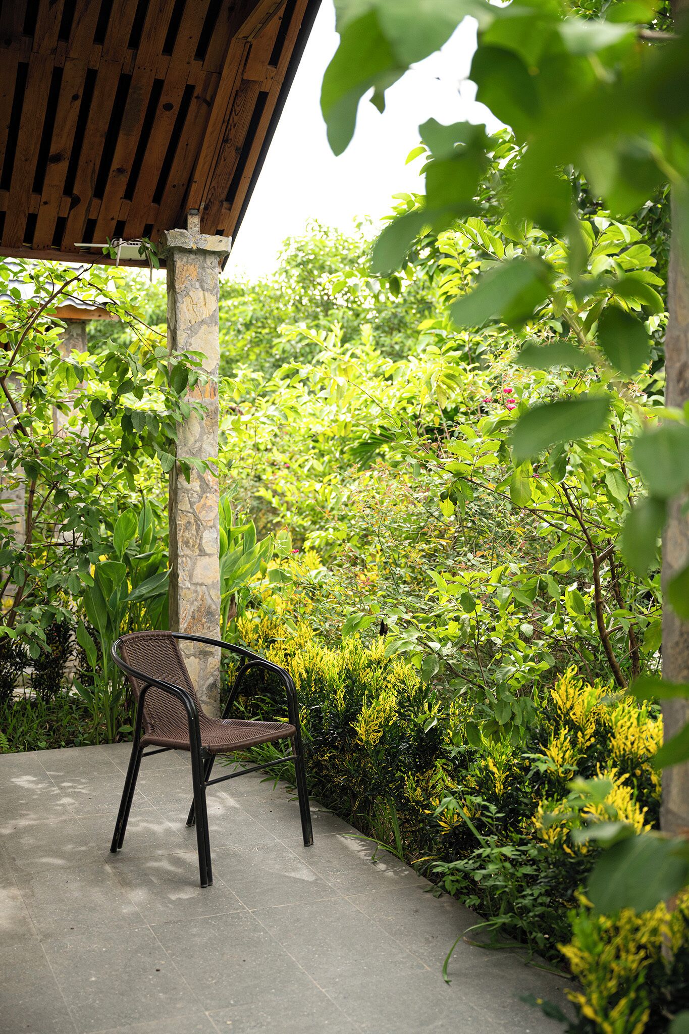 A covered outdoor seating area with a chair, surrounded by lush greenery and a stone pillar.