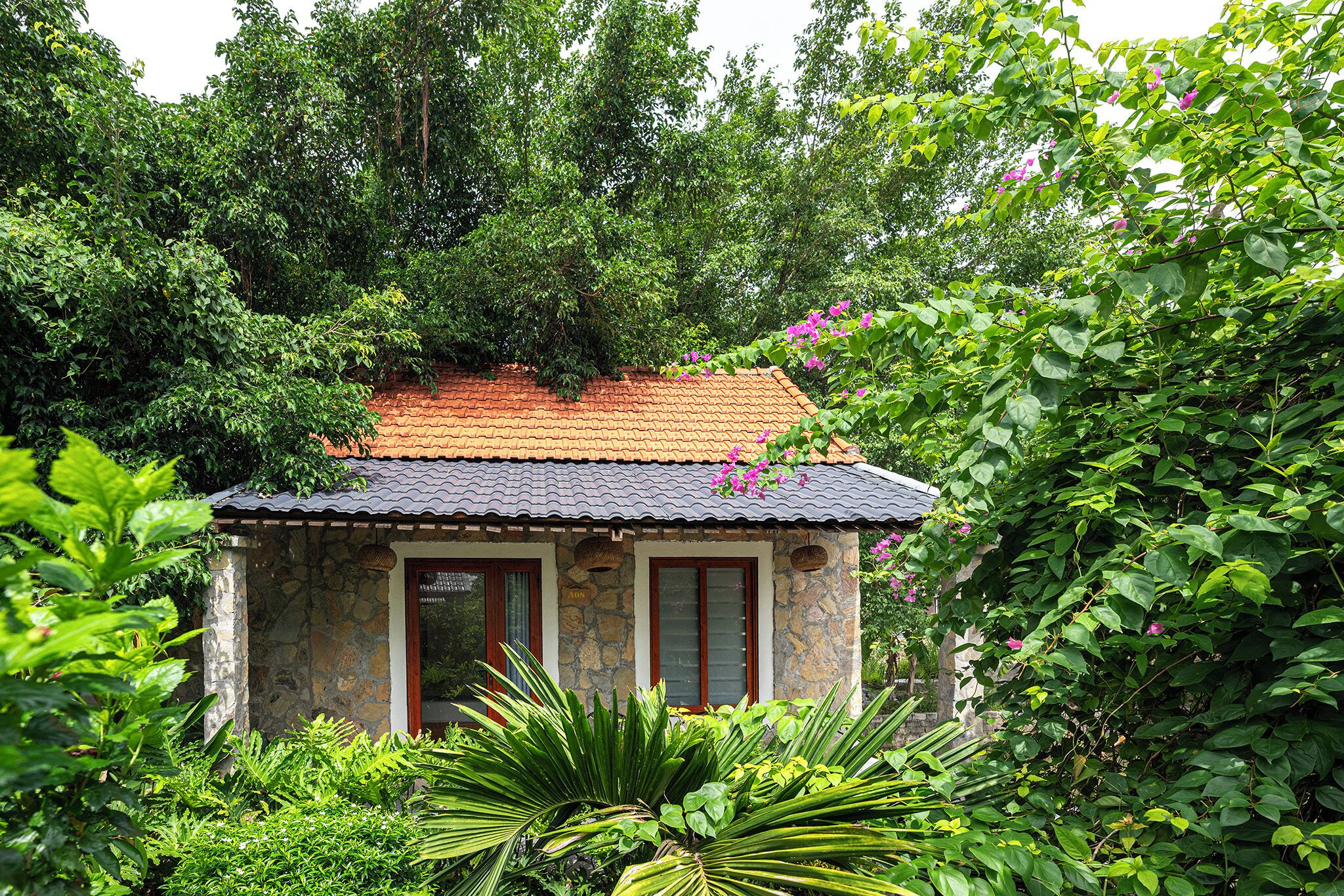 A small stone building with a tiled roof, surrounded by lush greenery and flowering plants.