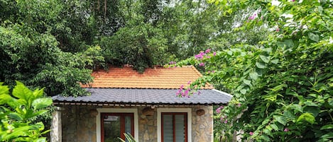 A small stone building with a tiled roof, surrounded by lush greenery and flowering plants.
