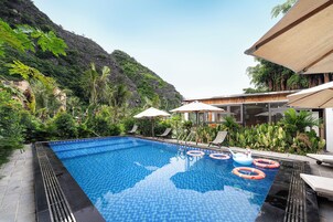 Una piscina con sillones de descanso y sombrillas, rodeada de exuberante vegetación y montañas al fondo.