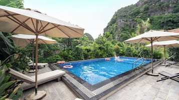 Una piscina con sillas de descanso y sombrillas, rodeada de abundante vegetación y montañas al fondo.