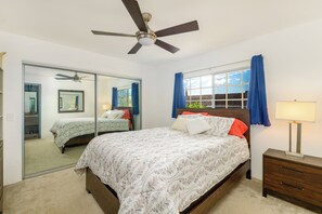 A bedroom with a bed, a ceiling fan, a mirror, a window with curtains, and a nightstand with a lamp.