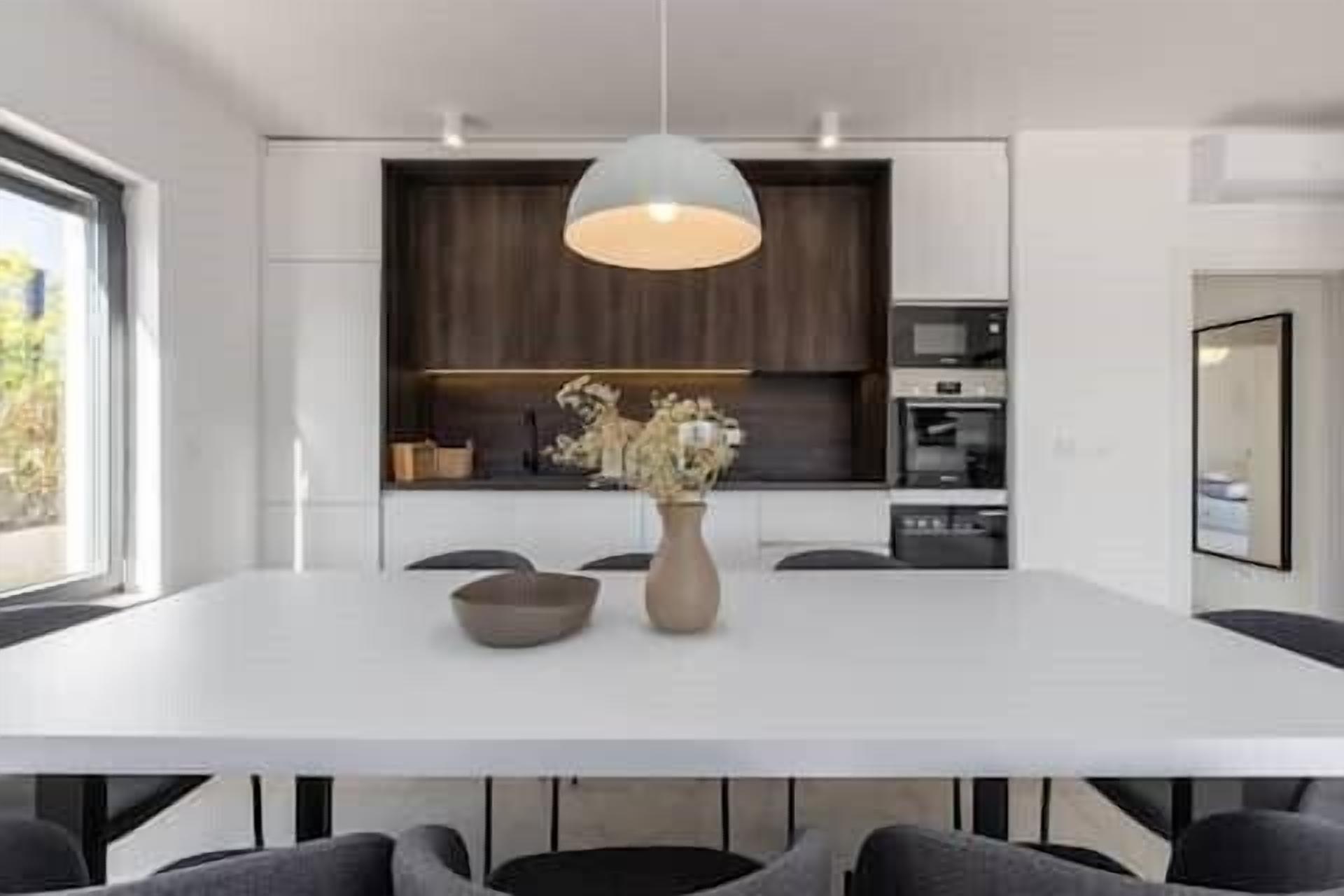 Un comedor moderno con una mesa blanca, sillas oscuras, un florero con flores y una lámpara colgante.