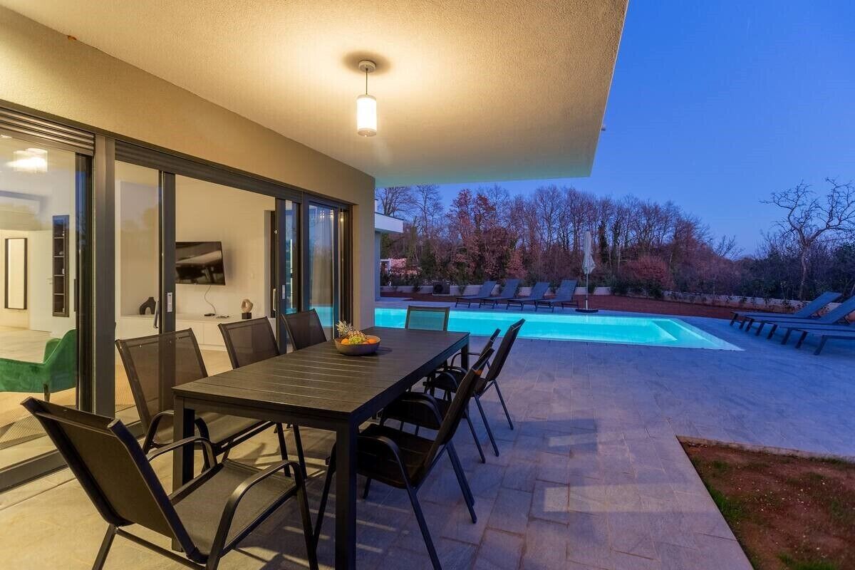 An outdoor patio with a dining table and chairs, a swimming pool, and a view of trees.