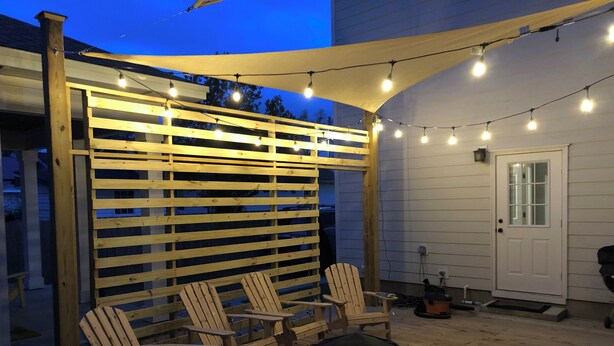 Um deck de madeira com cadeiras Adirondack e uma mesa redonda, sob uma pérgola com luzes de corda.