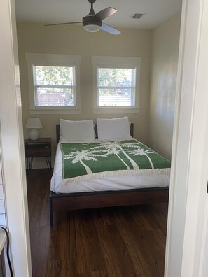 Un dormitorio con cama, dos ventanas, ventilador de techo y mesita de noche con lámpara.