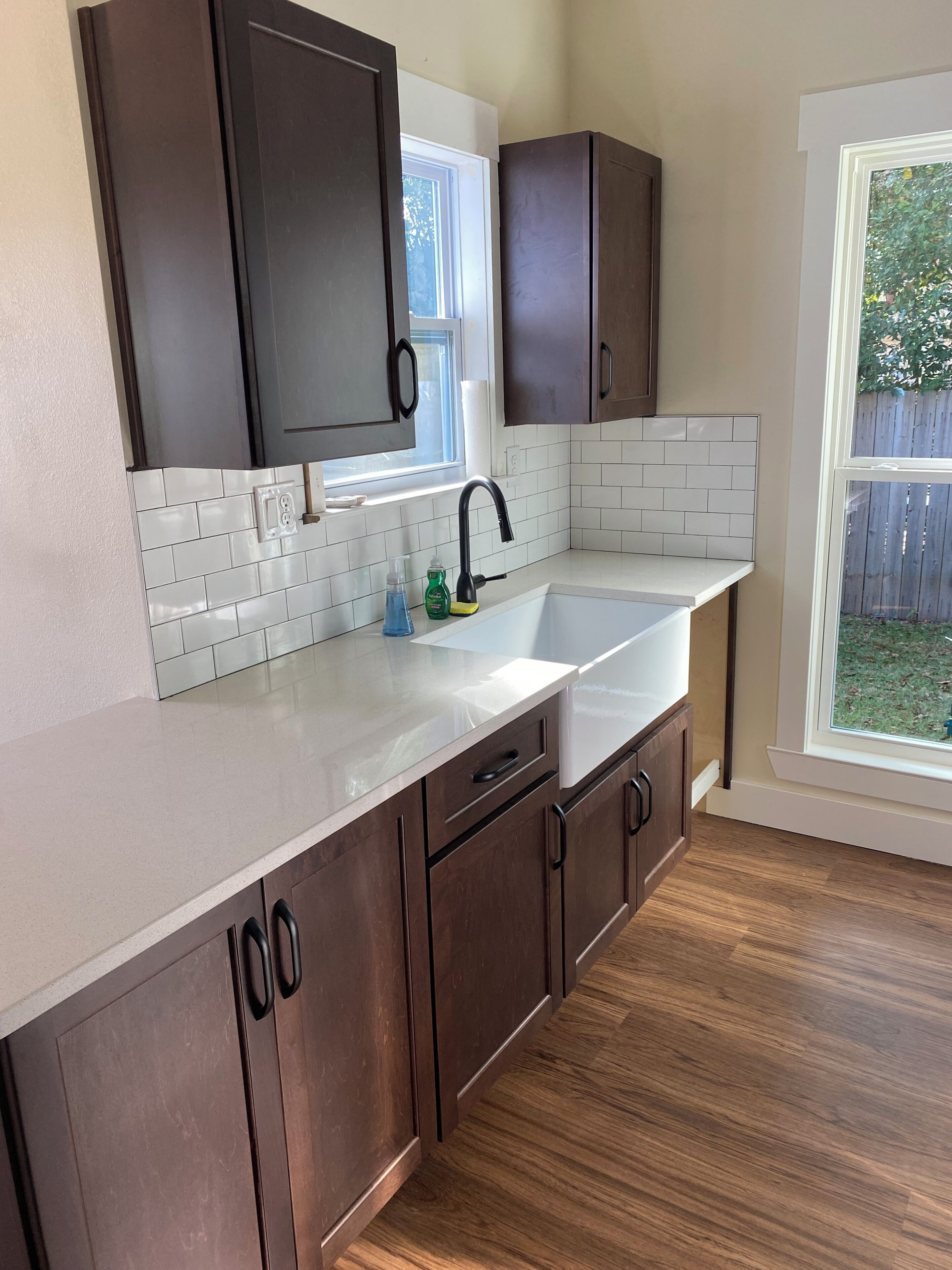 A kitchen with dark wooden cabinets, a white countertop, a sink, and a window with a view of the outdoors.