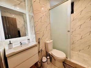 A bathroom with a marble wall, a toilet, a sink with a mirror, and a shower area. - 🌟 Luxury & Designer 2-Bedroom Apartment — Millennium Boulevard, Oran (Oran)
