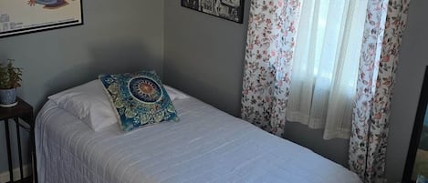 Ett sovrum med en säng, en inramad fotokollage, ett fönster med blommiga gardiner och en randig matta.