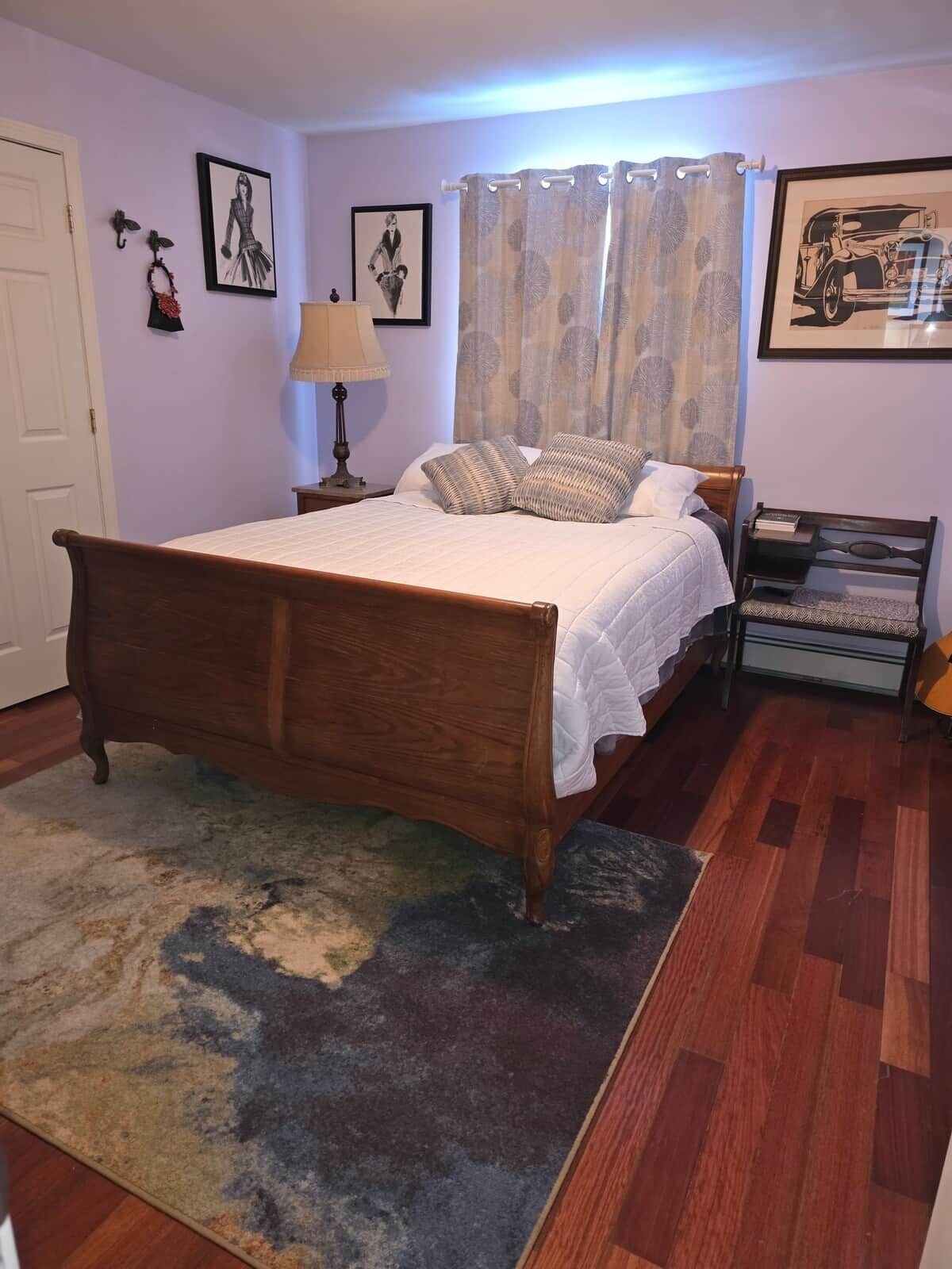 A bedroom with a wooden bed, a patterned rug, a nightstand with a lamp, and a small shelf with books.