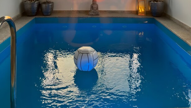 Una piscina con una estatua de Buda y plantas en macetas, vista al atardecer a través de un gran ventanal y un farol decorativo.