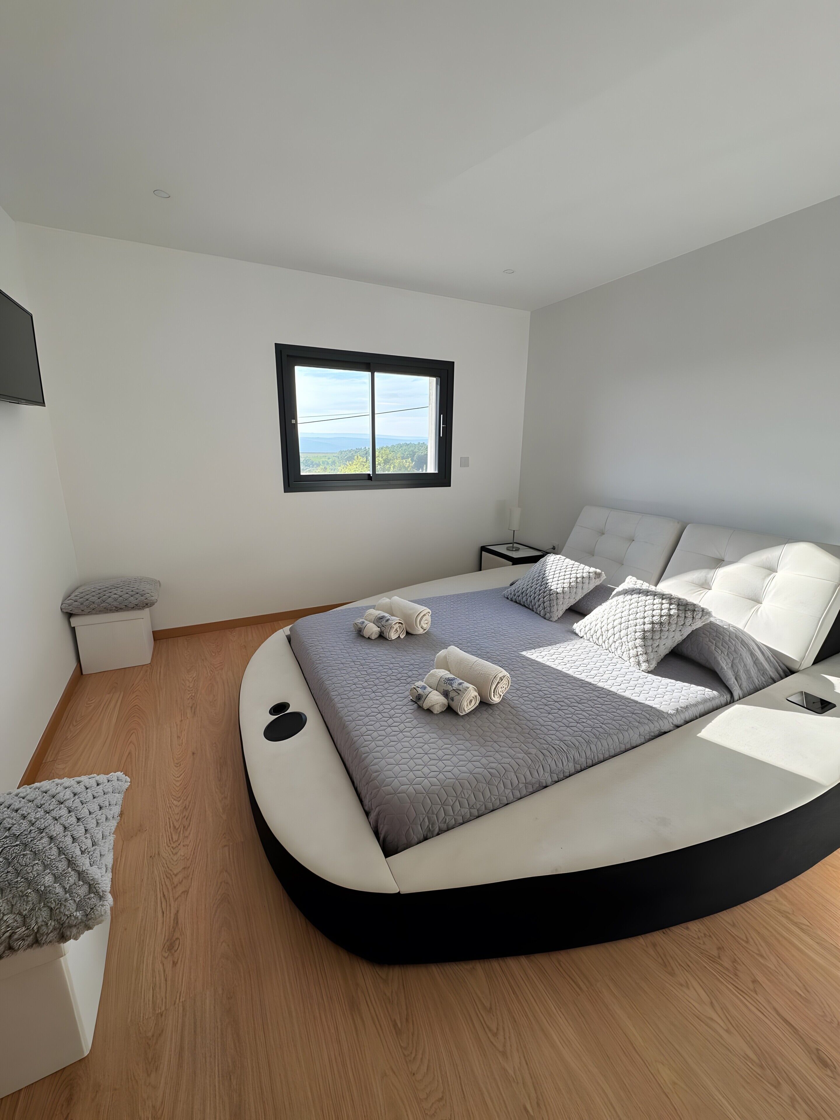 A modern bedroom with a curved bed, a view through a window, and a bedside table.