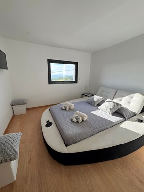 A modern bedroom with a curved bed, a view through a window, and a bedside table.