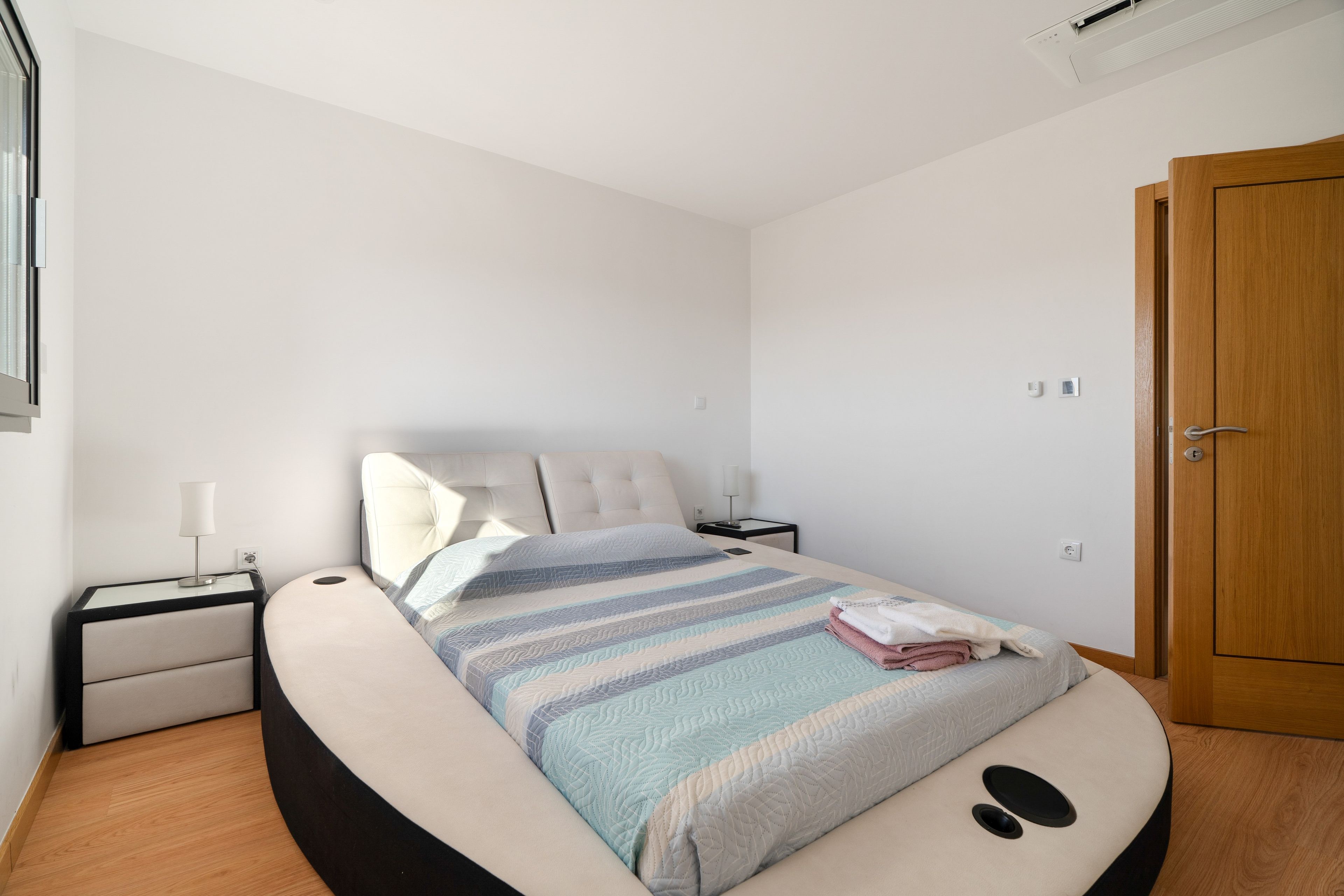 Quarto com cama, mesa de cabeceira, abajur, porta e janela.