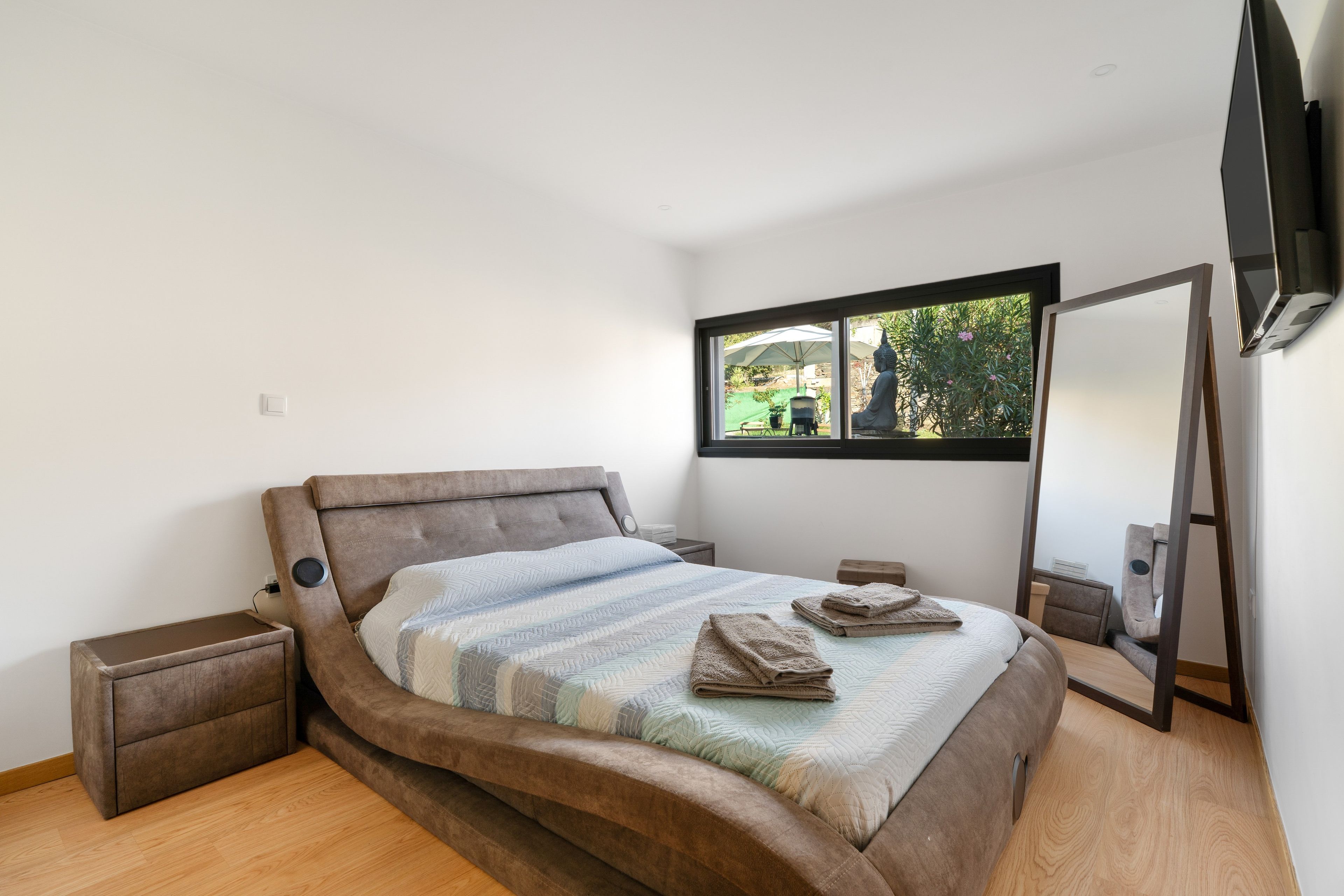 Un dormitorio moderno con una cama grande, una mesita de noche, un mueble auxiliar, una ventana con vistas a zonas verdes y un televisor de pared.