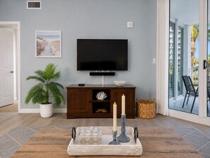 A living room with a flat-screen TV, a potted plant, a decorative tray with candles, and a large window with a view of palm trees and buildings. - Cozy Coastal Escape, Resort Pool + Walk to Beach (Destin)