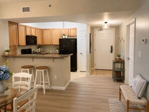 A modern kitchen with wooden cabinets, a black refrigerator, and a built-in microwave. There is a small island with stools and a vase of flowers. - Cozy Coastal Escape, Resort Pool + Walk to Beach (Destin)