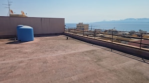Una terrazza sul tetto con un serbatoio d'acqua blu, una ringhiera metallica e vista sul mare e sulle montagne lontane.
