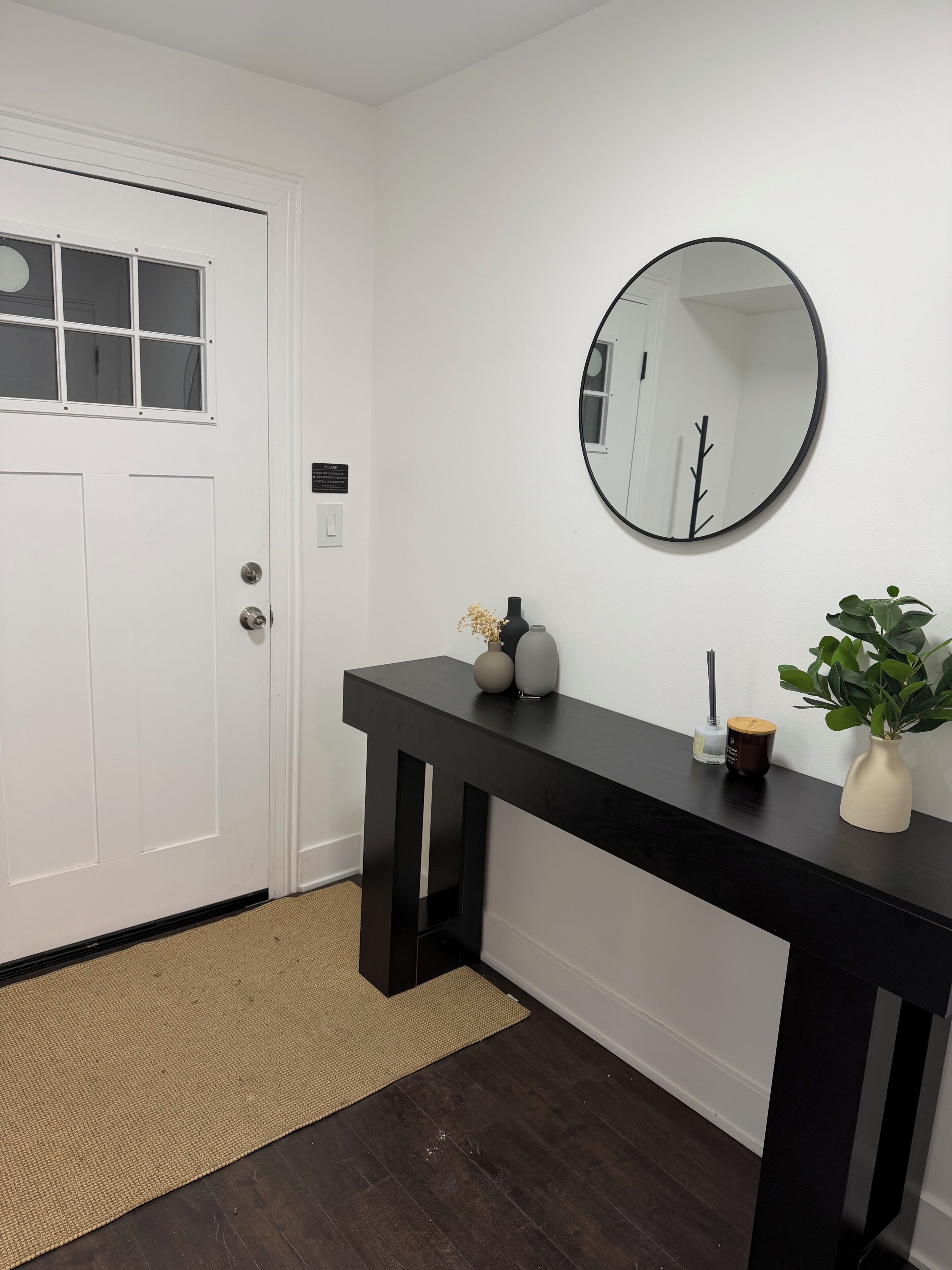 A hallway with a white door, a black console table with decorative items, a round mirror, and a woven mat on the floor.
