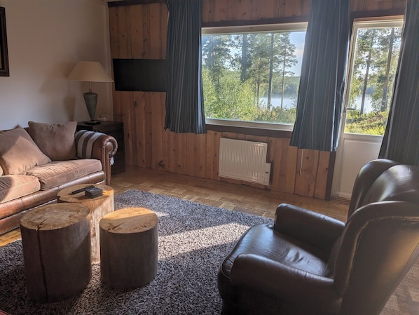 A cozy living room with a wooden interior, a leather sofa, a coffee table, and a view of a lake through a large window.