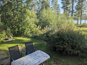 A wooden table with two chairs in a grassy area with dense foliage and a view of a lake. - Vrbo Property (Ytterhogdal)