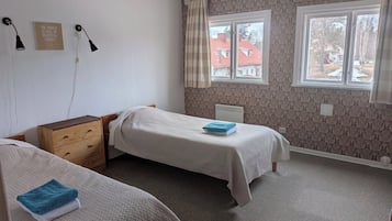 Ett sovrum med två enkelsängar, en byrå i trä, ett element och ett fönster med utsikt över byggnaderna utanför.