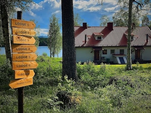 A wooden signpost with directions to various lodges and a beach, with a house and trees in the background. - Vrbo Property (Ytterhogdal)