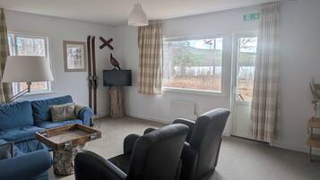 Ett rum med en blå soffa, ett soffbord i trä, två svarta fåtöljer, en tv på ett träställ, en fågelskulptur och ett fönster med gardiner.