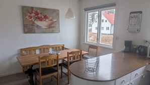 Un coin repas avec une table et des chaises en bois, un meuble blanc avec des tiroirs, une fenêtre donnant sur des immeubles et un tableau accroché au mur.