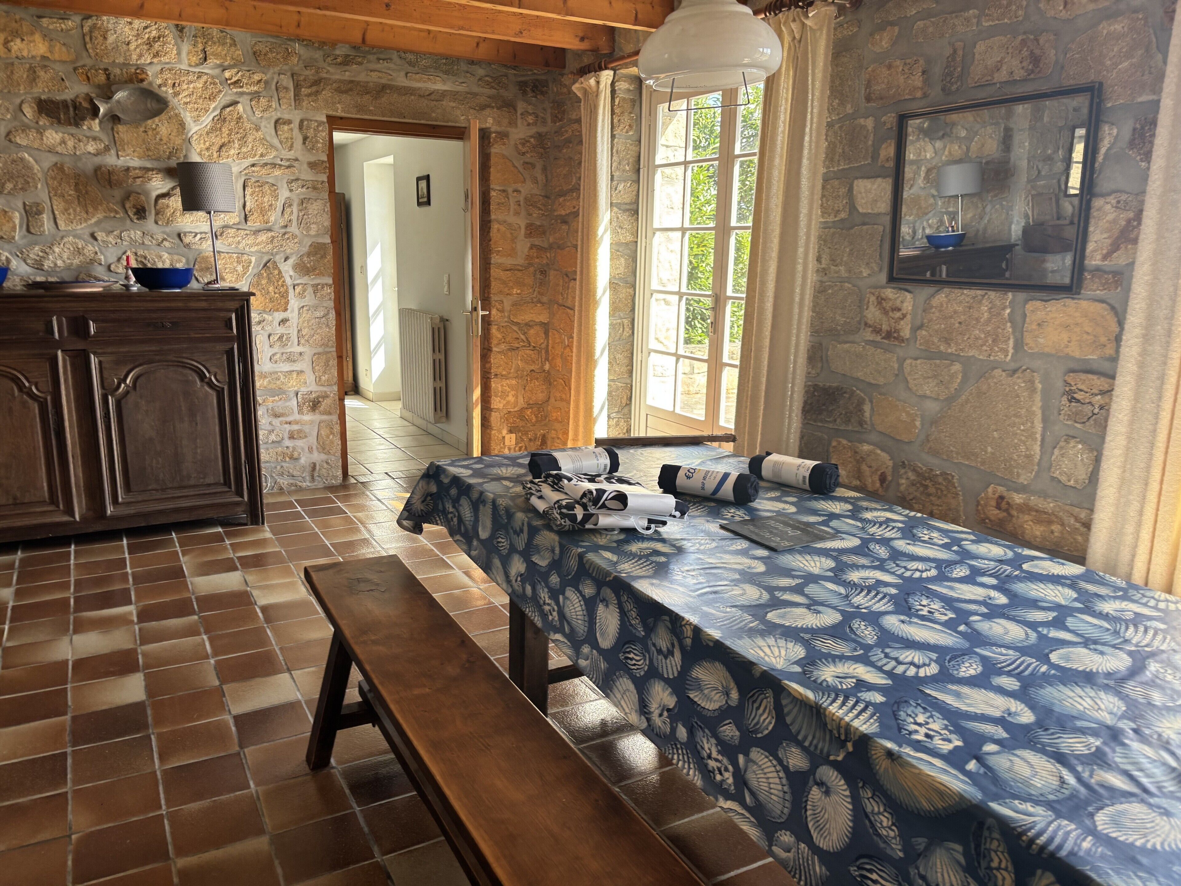 Una habitación con pared de piedra, un banco de madera, una mesa con mantel azul y una ventana con cortinas.